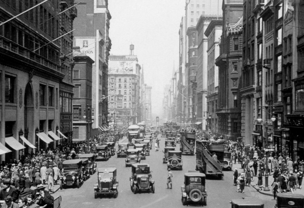 Historisches Schwarz-Weiß-Bild einer belebten Stadtstraße, bevölkert von Fußgängern und Autos.